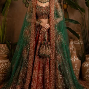 Emerald Green & Deep Red Bridal Lehenga – A Regal Expression of Indian Couture Make an unforgettable entrance in this Emerald Green & Deep Red Bridal Lehenga, an opulent designer ensemble created for brides and wedding guests who love traditional Indian craftsmanship with a luxurious, regal finish. Rich deep red tones are beautifully paired with jewel-like emerald green, while intricate gold embellishment brings depth, dimension, and grandeur to the complete look. This statement lehenga ensemble captures the essence of traditional Indian bridal fashion through its richly embroidered skirt, embellished blouse, and dramatic sheer green dupatta. Every element works together to create a sophisticated silhouette that feels festive, luxurious, and timeless. The deep red lehenga provides a classic bridal foundation, while the emerald green dupatta introduces a striking contrast. The extensive gold-toned embroidery throughout the outfit creates a luminous finish that makes the ensemble especially suited to grand celebrations, wedding ceremonies, receptions, sangeet nights, and formal festive occasions. The overall styling has a distinctly royal character, making this outfit an ideal choice for a bride or wedding guest who wants her outfit to feel rich, memorable, and beautifully detailed. Rich Deep Red and Emerald Green Colour Combination The combination of deep red, emerald green, and antique gold creates one of the most luxurious colour stories in Indian occasion wear. Deep red has long been associated with celebration, tradition, love, and bridal elegance. In this ensemble, the red base gives the lehenga visual richness while allowing the gold embroidery to stand out beautifully. The emerald green dupatta adds a sophisticated jewel-toned contrast. Rather than using green as the primary colour throughout the outfit, the design introduces it through the sheer dupatta, creating a layered appearance that feels dramatic without becoming overwhelming. Gold embroidery connects both colours and creates visual harmony between the red lehenga and green dupatta. This colour palette is particularly beautiful under warm wedding lighting, where the gold embellishment can catch the light and create a radiant appearance. Heavily Embroidered Lehenga Skirt The lehenga skirt is the centrepiece of this designer ensemble. Its deep red base is covered with intricate gold-toned embroidery arranged in elaborate traditional-inspired patterns. The embroidery creates an ornate surface that gives the skirt a rich couture appearance. The detailing continues across the length of the skirt, creating vertical visual movement and making the silhouette appear graceful and elongated. The lower hem features an especially elaborate decorative border. Multiple layers of embroidery and embellishment define the hemline and add weight and grandeur to the overall design. As the lehenga moves, the detailed embroidery catches the light, creating subtle flashes of gold throughout the skirt. This makes the outfit particularly beautiful for bridal portraits, wedding entrances, stage photographs, and evening celebrations. Statement Embellished Blouse The matching blouse has been designed to complement the richness of the lehenga without competing with it. The deep red base is decorated with intricate gold embroidery, creating a beautifully coordinated appearance from the waist upward. The fitted silhouette provides structure and definition while allowing the ornate detailing to remain the visual focus. The blouse is particularly well suited to traditional bridal jewellery. A statement choker, layered necklace, traditional earrings, maang tikka, and bangles can all be incorporated into the styling. The detailed sleeves and neckline also contribute to the overall couture appearance of the ensemble. For a customised order, blouse fitting, neckline preferences, sleeve styling, and other measurements can be discussed with the boutique depending on the selected design and customisation requirements. Sheer Emerald Green Designer Dupatta The emerald green dupatta is one of the most distinctive elements of this outfit. Crafted in a sheer-looking fabric appearance, the dupatta creates an elegant translucent layer over the richly embroidered lehenga. The transparency allows the deep red and gold embroidery underneath to remain visible while adding a beautiful green dimension to the overall look. The dupatta is heavily bordered with intricate gold detailing. The border frames the fabric beautifully and creates a strong visual outline when draped around the body. Large ornamental motifs are distributed across the dupatta, adding drama and movement. These motifs resemble radiant floral or sunburst-inspired patterns and create eye-catching focal points throughout the fabric. The combination of the transparent green layer and ornate gold embellishment gives the dupatta a luxurious, almost heirloom-inspired quality. Traditional Indian Bridal Craftsmanship The design reflects the beauty of traditional Indian bridal craftsmanship, where intricate embroidery, ornate borders, rich colours, and layered textiles come together to create a statement ensemble. Rather than relying on one type of embellishment, the outfit uses multiple levels of detailing. The skirt contains dense embroidery, the blouse carries coordinated embellishment, and the dupatta introduces a combination of sheer fabric, ornate borders, and statement motifs. This layered approach creates depth and richness. The result is an outfit that looks detailed from a distance while revealing even more intricate craftsmanship when viewed closely. Perfect for Bridal Celebrations This designer lehenga is an excellent choice for women looking for a luxurious outfit for their wedding celebrations. It can be styled for: Wedding ceremonies Bridal receptions Sangeet functions Engagement ceremonies Mehendi celebrations Family wedding events Cocktail receptions Festive celebrations Luxury destination weddings Formal cultural events Wedding guest dressing Bridesmaid styling For a bride, the deep red colour maintains a traditional bridal character, while the emerald green dupatta provides a distinctive contemporary contrast. For a wedding guest or family member, the ensemble offers the grandeur of bridal-inspired fashion while allowing personal styling to determine how dramatic or understated the final appearance becomes. A Regal Silhouette for the Modern Bride The silhouette of the lehenga creates a classic feminine shape while the embroidery gives it a modern couture finish. The full skirt creates beautiful volume and movement. When paired with the fitted blouse, it produces a balanced silhouette that works especially well for formal Indian celebrations. The long flowing dupatta further enhances the silhouette by creating a graceful frame around the body. This combination is particularly effective for photographs because the outfit has several distinct layers that create depth. The lehenga can be draped traditionally over one shoulder or styled more dramatically around the arms depending on the desired appearance. Gold Embroidery That Catches the Light Gold is an essential element of this ensemble. The embroidery is distributed throughout the skirt, blouse, and dupatta, ensuring that the outfit maintains a consistent visual language. Under wedding lights, the gold detailing can create a beautiful luminous effect. This makes the lehenga especially suitable for evening functions, where the combination of rich red, emerald green, and gold can appear particularly dramatic. The detailed borders also help define the silhouette and create visual structure around the hemline and dupatta. For brides who want a richly embellished outfit that photographs beautifully, this type of detailing can create a striking result. Bridal Jewellery Styling This lehenga offers numerous jewellery styling possibilities. For a traditional bridal appearance, pair it with a heavy gold or antique-finish choker, matching earrings, a maang tikka, and layered necklaces. For a more regal look, add a long rani haar beneath the choker. The deep red and emerald combination works beautifully with traditional jewellery featuring green and gold accents. For a contemporary bride, you can reduce the number of jewellery pieces and focus on one statement choker with large earrings. Bangles can be stacked in complementary shades to enhance the traditional character of the outfit. A traditional bridal nath can also be added for a more ceremonial appearance. Hairstyle Suggestions A sleek traditional bun is an excellent hairstyle for this lehenga. A centre-parted bun decorated with flowers can create a classic bridal appearance while keeping the neckline and jewellery visible. For a royal-inspired look, consider a low bun with traditional hair accessories. If you prefer a more contemporary style, soft waves can be worn with the dupatta draped over one shoulder. A neatly styled bun is particularly useful when wearing a maang tikka because it keeps the overall bridal look balanced. Makeup Suggestions The rich colours of this lehenga pair beautifully with warm and glamorous makeup. A softly defined eye with gold or bronze tones can complement the embroidery. A deeper liner and carefully defined brows can enhance the eyes without overpowering the jewellery. For the complexion, a luminous finish can complement the richness of the outfit. Warm blush, subtle contouring, and a classic red, berry, or deep rose lip can complete the bridal appearance. For a softer daytime celebration, choose a muted rose or nude lip and lighter eye makeup. For an evening reception, a deeper lip and more defined eye look can create a dramatic finish. Perfect for Sangeet and Reception Nights Although the colour palette has a traditional bridal character, this lehenga can be styled beautifully for modern sangeet and reception celebrations. For a sangeet, pair it with statement earrings, a sleek hairstyle, and glamorous makeup. The emerald green dupatta can be draped in a way that allows maximum movement while dancing and celebrating. For a reception, style the outfit with sophisticated jewellery, an elegant clutch, and a polished hairstyle. The gold embroidery ensures that the ensemble remains glamorous enough for a formal evening event. Destination Wedding Styling The rich colours and ornate embroidery of this lehenga can also make a beautiful statement at a destination wedding. Whether the celebration takes place in a heritage palace, luxury hotel, resort, or traditional wedding venue, the combination of red, green, and gold photographs beautifully against warm architectural backgrounds. For a destination wedding, the dupatta can be styled in a lighter, more relaxed drape while keeping the embroidered border visible. Statement jewellery and a structured clutch can complete the look. An Heirloom-Inspired Design The colour combination and detailed embroidery give this ensemble an heirloom-inspired quality. Traditional bridal outfits are often cherished because they become connected with important memories. A carefully crafted lehenga can become part of a family's visual history, appearing in wedding photographs and being remembered long after the celebration. This design combines classic colours with an elaborate embroidery aesthetic, making it suitable for women who appreciate timeless Indian fashion. Its traditional character means it can remain visually relevant even as fashion trends change. Custom-Made Bridal Experience At Maharani Designer Boutique, selected bridal and occasion-wear designs can be developed with individual requirements in mind. For customers choosing a custom-made or made-to-order outfit, measurements, fitting requirements, event dates, and selected styling details can be discussed before production. Depending on the design, customers may be able to discuss elements such as blouse fitting, neckline preferences, sleeve requirements, dupatta styling, and other finishing details. Because heavily embroidered bridal outfits require careful craftsmanship and production time, customers are encouraged to share their wedding date in advance. The boutique can then advise on the appropriate production timeline based on the selected design and requirements. Why Choose a Designer Bridal Lehenga? A bridal lehenga is more than an outfit for one day. It is an expression of personal style and an important part of the wedding experience. Choosing a designer lehenga allows you to focus on details such as colour harmony, embroidery placement, silhouette, finishing, and overall styling. This emerald green and deep red ensemble has been designed to create a complete look rather than simply a decorated skirt and blouse. The coordinated colours, borders, embroidery, and dupatta motifs work together to produce a cohesive appearance. This makes the ensemble particularly suitable for women who want their bridal clothing to feel thoughtfully designed from every angle. Ideal for Indian and International Weddings Indian weddings are celebrated around the world, and contemporary brides often look for authentic Indian craftsmanship combined with modern styling. This lehenga can be an excellent option for customers in India as well as Indian families celebrating weddings internationally. Whether your wedding takes place in Jalandhar, Delhi, Chandigarh, Mumbai, Canada, the USA, the UK, Australia, New Zealand, or another destination, the rich colour palette can create a striking bridal appearance. Maharani Designer Boutique serves customers looking for premium Indian ethnic wear and designer wedding outfits with shipping options available across India and internationally. Styling the Emerald Dupatta The emerald green dupatta can be styled in several ways. For a traditional bridal drape, place it over the head and allow the embroidered border to frame the face. For a contemporary style, drape it over one shoulder and let the fabric fall naturally down the back. Another option is to create a structured shoulder drape that showcases the ornate border while keeping the blouse clearly visible. The transparency of the fabric means that the red lehenga continues to remain visible underneath, creating a layered colour effect. This is one of the key reasons the green dupatta gives the outfit such a distinctive appearance. The Beauty of Contrasting Colours One of the strongest design elements of this ensemble is the use of contrasting colours. The deep red provides warmth and richness. The emerald green introduces freshness and jewel-toned depth. Gold connects the two colours and adds luminosity. This three-colour combination creates a sophisticated visual balance. None of the colours needs to dominate completely because they each serve a specific purpose within the overall design. The result is an outfit that feels luxurious, traditional, and dramatic while still maintaining a carefully controlled aesthetic. Wedding Guest and Family Styling While the ensemble can certainly be styled as bridal wear, it can also be considered for close family members attending an elaborate wedding. A bride's sister, cousin, sister-in-law, mother, or close family member can style the outfit according to the occasion and level of jewellery desired. For a family wedding, reducing the jewellery and choosing a simpler hairstyle can create a sophisticated formal appearance. For a bridal reception, more elaborate jewellery can transform the same ensemble into a dramatic statement look. Photography-Friendly Design A well-designed wedding outfit should look beautiful both in person and in photographs. This lehenga features several elements that naturally create visual interest for photography. The deep red skirt provides a rich foundation. The gold embroidery creates texture and highlights. The emerald dupatta adds a contrasting layer. The large motifs on the dupatta create additional focal points. The ornate hem gives the skirt definition when photographed from a distance. Together, these elements create a complete visual story that can work beautifully in bridal portraits, couple photographs, family photographs, and wedding ceremony images. Care and Handling Because this is an intricately embellished occasion-wear ensemble, careful handling is recommended. Avoid excessive friction against rough surfaces and store the outfit in a clean, dry garment bag when it is not being worn. After the event, professional cleaning appropriate to the fabric and embellishment is recommended. The embroidered components should be handled carefully to help preserve their detailing and finish. Before cleaning, always confirm the recommended care method for the actual fabric and embellishment used in the final garment. Order Your Designer Bridal Lehenga If you are searching for a red and green bridal lehenga, designer bridal lehenga online, heavy embroidered lehenga, wedding lehenga, or luxury Indian bridal outfit, this ensemble offers a beautifully regal choice. Its deep red base, emerald green sheer dupatta, extensive gold embroidery, statement borders, and traditional-inspired motifs create an unforgettable occasion-wear look. Whether you are planning your wedding, preparing for a reception, attending a sangeet, or looking for an elegant family wedding outfit, this designer lehenga provides a luxurious foundation for your personal styling. Discover the latest designer Indian wear collection at Maharani Designer Boutique and create a wedding look that reflects elegance, tradition, craftsmanship, and individuality. For availability, pricing, measurements, customisation, production timelines, and shipping information, contact the boutique directly. Product Highlights Deep red designer lehenga Emerald green sheer-style dupatta Heavy gold-toned embroidery Intricately embellished blouse Elaborately decorated lehenga hem Detailed dupatta border Statement embroidered motifs Regal red, green, and gold colour combination Suitable for brides and wedding celebrations Ideal for sangeet and reception functions Suitable for engagement and festive occasions Custom-made options may be available Made-to-order assistance available depending on requirements PAN India and worldwide shipping Red green bridal lehenga, red and green lehenga, designer bridal lehenga, bridal lehenga online, heavy bridal lehenga, designer lehenga online, wedding lehenga, bridal lehenga India, embroidered bridal lehenga, heavy embroidered lehenga, red bridal lehenga, emerald green dupatta lehenga, luxury bridal lehenga, designer wedding lehenga, Indian bridal wear, Indian wedding lehenga, bridal outfit online, lehenga for wedding, lehenga for reception, sangeet lehenga, engagement lehenga, traditional bridal lehenga, royal bridal lehenga, couture bridal lehenga, custom made bridal lehenga, designer boutique lehenga, bridal lehenga Jalandhar, Maharani Designer Boutique, designer ethnic wear India.