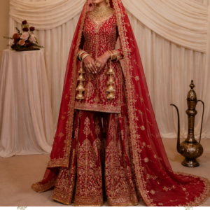 Ruhani Riwaayat – Regal Red Bridal Sharara Suit Make your most cherished celebrations unforgettable with Ruhani Riwaayat, a magnificent red bridal sharara suit designed by Maharani Designer Boutique. Created for the modern bride who treasures traditional Indian craftsmanship, this luxurious ensemble combines a rich red colour palette, intricate golden embroidery, a beautifully detailed short kurta, an ornate sharara, and a statement dupatta that completes the bridal silhouette. The deep red base gives the outfit a classic bridal character, while the elaborate golden embroidery creates a luxurious and regal appearance. Every element of the ensemble works together to create a harmonious traditional look, from the richly decorated neckline and bodice to the embroidered hemline, flowing sharara panels, and wide dupatta border. Ruhani Riwaayat is designed for women who want to embrace the timeless beauty of Indian wedding fashion while maintaining an elegant and sophisticated appearance. It can be styled for bridal celebrations, engagement ceremonies, wedding festivities, reception events, family weddings, sangeet functions, and other grand occasions. The outfit beautifully represents the charm of traditional South Asian occasion wear. Its red-and-gold combination instantly evokes the grandeur of Indian bridal fashion, while the sharara silhouette adds movement, volume, and elegance. Whether you are the bride, a close family member, or a woman looking for an exceptionally luxurious wedding outfit, Ruhani Riwaayat offers a statement-making choice. A Timeless Red Bridal Colour Red has always held a special place in Indian bridal fashion. It represents celebration, beauty, confidence, tradition, and grandeur. The rich red tone of Ruhani Riwaayat gives the ensemble an unmistakably festive personality. Rather than relying on multiple contrasting colours, the design uses red as the dominant base and allows the golden embroidery to become the primary decorative element. This creates a sophisticated red-and-gold combination that works beautifully for bridal photography. Under warm lighting, the golden embroidery catches attention against the deep red base, creating a luxurious visual effect. The colour is particularly suitable for: Bridal ceremonies Wedding receptions Sangeet celebrations Engagement functions Roka ceremonies Family weddings Festive celebrations Traditional wedding portraits Formal evening events For brides who love classic Indian wedding colours, this red sharara suit offers an elegant interpretation of traditional bridal dressing. Bridal Designer Lehenga , MDB 024848 , MDB 026342 , MDB 027210229, MDB 027210275, Intricate Golden Embroidery The most striking feature of Ruhani Riwaayat is its elaborate golden embroidery. The embroidery covers significant portions of the outfit while maintaining a balanced pattern. Floral, ornamental, and traditional-inspired motifs are distributed across the kurta and sharara, creating an ornate appearance. The detailing around the neckline draws attention toward the face. The embroidery continues across the bodice, sleeves, lower hem, and sharara panels. The border work adds definition to each garment and gives the entire outfit a structured finish. Golden embroidery against red creates a classic combination that has remained popular in Indian occasion wear for generations. The richness of the embroidery also makes the outfit particularly suitable for formal photographs and evening celebrations. Elegant Short Kurta The kurta features a refined short silhouette designed to complement the voluminous sharara. The neckline is beautifully decorated with dense golden embroidery. The embroidered neckline creates a strong focal point around the face and allows the wearer to style the outfit with a statement choker, necklace, or traditional bridal jewellery. The front of the kurta features all-over ornamental embroidery. The sleeves are also richly decorated, maintaining continuity between the upper and lower portions of the outfit. The hem features a prominent embroidered border that separates the kurta from the sharara while keeping the overall look cohesive. The shorter kurta creates a balanced proportion with the wide sharara silhouette. Statement Sharara The sharara is the defining element of the ensemble. Its wide, flowing silhouette creates beautiful movement while walking. Each panel is richly decorated with golden motifs. The embroidery is arranged vertically and across the lower sections, giving the sharara a luxurious appearance. The broad embroidered borders at the hem provide additional structure and definition. When the wearer moves, the wide sharara creates graceful folds that reveal the embroidery from different angles. This makes the outfit particularly attractive for wedding entrances, sangeet performances, bridal portraits, and celebration photographs. The sharara silhouette also offers a beautiful alternative to the traditional lehenga for women who want a distinctive wedding outfit. Ornate Sharara Borders The lower border of the sharara features intricate golden detailing. The border creates a strong visual foundation for the entire outfit. Its elaborate design complements the embroidery on the kurta and dupatta. The layered appearance of the border adds richness without taking attention away from the overall silhouette. The combination of vertical motifs and horizontal borders creates a balanced pattern. This detailed finishing makes the sharara look especially beautiful in full-length photographs. Regal Dupatta The dupatta completes the ensemble with an elegant flowing silhouette. The red dupatta coordinates perfectly with the kurta and sharara. Its outer border features rich golden embroidery. Small motifs are distributed across the body of the dupatta, creating a sophisticated balance between the central fabric and the heavy border. The generous length allows the dupatta to be styled traditionally over the head or draped across the shoulders. For bridal styling, the dupatta can be placed over the head to create a traditional bridal appearance. For a reception or sangeet, it can be draped over one shoulder for a more contemporary silhouette. Beautiful Dupatta Border The heavily embroidered dupatta border is one of the most important design elements. It frames the red fabric with a rich golden edge. When placed over the head, the border beautifully surrounds the face. When draped over the shoulder, the embroidered edge creates a statement vertical line. When allowed to trail behind the wearer, it adds drama and movement to bridal photographs. The border also visually connects the dupatta with the embroidered borders of the kurta and sharara. Perfect Bridal Sharara Suit Ruhani Riwaayat is especially suitable for brides searching for an alternative to the conventional bridal lehenga. A sharara provides a different silhouette while maintaining the grandeur expected from bridal clothing. The wide trousers create movement and volume. The short kurta balances the proportions. The flowing dupatta adds the traditional bridal finishing layer. Together, these three pieces create a complete and luxurious bridal ensemble. The red colour further enhances its bridal character. Ideal for Wedding Functions This ensemble can be styled for different wedding celebrations. Engagement For an engagement ceremony, style it with a statement necklace, earrings, and elegant heels. Sangeet For a sangeet, the sharara silhouette provides beautiful movement and is ideal for dancing and photographs. Wedding For the wedding ceremony, wear the dupatta over the head and pair it with traditional bridal jewellery. Reception For a reception, style the dupatta over one shoulder and choose statement earrings with a sophisticated hairstyle. Family Wedding The ensemble is also suitable for close family members who want a luxurious traditional appearance. Bridal Jewellery Styling Ruhani Riwaayat works beautifully with traditional Indian jewellery. For a classic bridal look, consider: Gold choker Long necklace Jhumka earrings Maang tikka Nath Bangles Hathphool Statement rings A layered jewellery combination can create a grand bridal appearance. For a more contemporary look, choose one statement choker and oversized earrings. Because the outfit already features rich embroidery, jewellery should be selected according to the desired level of grandeur. Traditional Bridal Makeup The rich red colour works beautifully with traditional bridal makeup. Consider warm gold or bronze eyeshadow, defined eyeliner, softly sculpted cheeks, and a classic red or deep berry lip. For a softer look, choose neutral eyes with a warm nude-pink lip. A luminous complexion can complement the golden embroidery beautifully. The final makeup style can be adjusted depending on the wedding function and time of day. Hairstyle Ideas A traditional bridal bun is an excellent choice when wearing the dupatta over the head. Decorate the bun with fresh flowers for a classic Indian bridal appearance. For a reception, soft waves or a low textured bun can create a more contemporary appearance. A centre-parted hairstyle works especially well with a maang tikka. If wearing a traditional nath, an elegant bun can help keep the overall bridal look balanced. Perfect for Bridal Photography The red-and-gold palette makes Ruhani Riwaayat particularly suitable for bridal photography. The embroidery creates visual texture. The flowing dupatta adds movement. The wide sharara provides volume. The deep red colour creates a striking contrast against neutral wedding décor. For full-length photographs, allow the sharara panels to spread naturally. For portraits, style the dupatta over the head and highlight the neckline and jewellery. For movement photographs, allow the dupatta to flow behind you. Traditional Yet Contemporary One of the strongest qualities of this design is its ability to combine traditional styling with a contemporary silhouette. The red-and-gold colour combination feels deeply rooted in Indian bridal culture. The sharara silhouette gives it a fashionable and distinctive character. The shorter kurta creates a modern proportion. The flowing dupatta preserves the traditional elegance expected from a formal Indian ensemble. This balance makes Ruhani Riwaayat suitable for women who want cultural richness without choosing a conventional lehenga. A Statement Outfit for the Modern Bride Modern brides increasingly look for outfits that express individuality. Ruhani Riwaayat offers a beautiful alternative to standard bridal silhouettes. The sharara allows freedom of movement while maintaining a dramatic appearance. The intricate embroidery ensures the outfit remains luxurious. The red colour maintains the traditional bridal character. The result is an ensemble that can feel both familiar and distinctive. Styling for Sangeet For a sangeet celebration, style the outfit with: Statement earrings Minimal necklace Comfortable embellished heels Soft waves Metallic clutch The sharara silhouette works particularly well for movement. Its wide panels create a beautiful effect while dancing or walking. For a coordinated family look, the outfit can also inspire complementary colours for bridesmaids or family members. Styling for Reception For a reception, create a glamorous appearance with: Statement diamond-style earrings Sleek hairstyle High heels Elegant clutch Defined eye makeup Soft berry lipstick Drape the dupatta across one shoulder instead of covering the head. This changes the character of the outfit from traditional bridal to contemporary evening wear. Styling for Roka or Engagement For a smaller celebration, keep the styling refined. Choose a delicate choker and matching earrings. Use soft makeup and a low bun. Allow the dupatta to fall naturally from one shoulder. The embroidery will provide sufficient visual detail without requiring excessive accessories. Versatility of the Ensemble Although designed with bridal aesthetics, Ruhani Riwaayat can be styled for several formal occasions. The three-piece design provides multiple styling possibilities. The dupatta can be worn differently. Jewellery can be changed. Hairstyles can be adjusted. Makeup can be softened or intensified. Footwear can transform the overall appearance. This versatility makes it a valuable addition to an occasion-wear wardrobe. Maharani Designer Boutique Maharani Designer Boutique specialises in elegant Indian ethnic wear designed for important celebrations. The brand's collections include designer Punjabi suits, bridal lehengas, sharara suits, Pakistani-inspired outfits, party wear suits, wedding dresses, and custom-made ethnic ensembles. Ruhani Riwaayat represents the luxurious side of the brand's occasion-wear aesthetic. The design combines traditional South Asian influences with detailed finishing and contemporary styling. Custom-Made Experience Depending on the specific order and design requirements, customisation can be discussed with Maharani Designer Boutique. Possible considerations may include: Personal measurements Fit adjustments Sleeve length Kurta length Sharara length Dupatta styling Colour preferences Embroidery variations Personal design requirements Customisation availability depends on the design and production requirements. For bridal orders, customers are encouraged to plan ahead and discuss their event date before placing an order. Suitable for Indian Weddings Worldwide Ruhani Riwaayat is suitable for South Asian weddings and celebrations across India and internationally. It can be styled for weddings in: India Canada USA UK Australia New Zealand Other international destinations The timeless red-and-gold colour combination works beautifully across different wedding themes and décor styles. Care and Storage Because this is an elaborately embroidered occasion-wear ensemble, careful handling is recommended. Store the outfit in a clean, breathable garment bag. Avoid placing heavy objects over the embroidered areas. Keep the garment away from excessive moisture. Fold the dupatta carefully to protect the embroidered border. Avoid contact with rough surfaces that could catch delicate embellishments. Professional cleaning is recommended where appropriate. For long-term storage, refold the garment periodically to help preserve its shape and embroidery. Always follow the specific care instructions supplied with the finished garment. Why Choose Ruhani Riwaayat? Choose this design if you are looking for: Red bridal sharara Red bridal sharara suit Red embroidered sharara Red wedding sharara Bridal sharara suit for women Designer sharara suit Bridal sharara online Red Pakistani sharara suit Red Punjabi sharara suit Red wedding outfit Red bridal outfit Red wedding wear for women Red engagement outfit Red sangeet outfit Red reception outfit Traditional Indian bridal wear Designer Indian ethnic wear Luxury bridal wear Embroidered bridal outfit Red and gold bridal outfit Red and gold sharara Golden embroidered sharara Wedding guest sharara Bridal party wear Designer wedding outfit Maharani Designer Boutique Designer boutique Jalandhar Bridal boutique Jalandhar Punjabi designer boutique Indian wedding clothing Luxury ethnic wear for women Product Details Product Name: Ruhani Riwaayat Brand: Maharani Designer Boutique Category: Bridal Sharara Suit Colour: Deep Red / Bridal Red Style: Traditional Luxury Ethnic Wear Top: Embroidered Short Kurta Bottom: Wide-Leg Embroidered Sharara Dupatta: Red Embroidered Dupatta with Ornate Border Embroidery: Intricate Golden Traditional-Inspired Embroidery Look: Regal, Traditional, Bridal & Festive Occasions: Wedding, Engagement, Sangeet, Reception, Roka, Festive Events & Formal Celebrations Customisation: Available depending on design and requirements Shipping: PAN India & Worldwide red bridal sharara, red bridal sharara suit, bridal sharara suit, red sharara suit, red wedding sharara, red embroidered sharara, designer sharara suit, bridal sharara online, red bridal outfit, red wedding outfit, red and gold bridal outfit, red and gold sharara, golden embroidered sharara, bridal sharara for wedding, bridal sharara for women, designer bridal sharara, luxury bridal sharara, Indian bridal sharara, Pakistani bridal sharara, Punjabi bridal sharara, red Pakistani sharara, red Punjabi sharara, Punjabi sharara suit, Pakistani sharara suit, sharara suit for wedding, wedding sharara for women, sangeet sharara, Bridal Sharara Suits engagement sharara, reception sharara, bridal party wear, wedding party wear, traditional bridal wear, Indian wedding wear, designer Indian ethnic wear, luxury ethnic wear for women, embroidered wedding outfit, embroidered bridal wear, red ethnic wear, red festive wear, wedding outfit for women, bridal outfit online, designer wedding outfit, bridal boutique, designer boutique Jalandhar, bridal boutique Jalandhar, Punjabi designer boutique, Maharani Designer Boutique, Maharani Designer bridal wear.