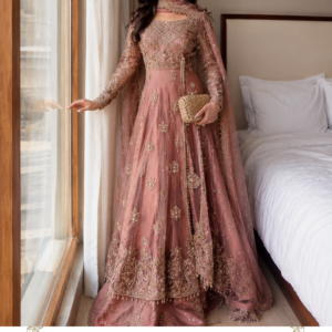 Gulnaar Rosé – A Romantic Statement in Dusty Rose Introducing Gulnaar Rosé, an elegant dusty rose pink designer ensemble from Maharani Designer Boutique, created for women who love graceful silhouettes, delicate pastel colours, and intricate traditional embellishment. This sophisticated three-piece ensemble brings together a beautifully embellished long Anarkali-style kurta, coordinated flared sharara trousers, and a sheer-looking matching dupatta decorated with ornate embroidery and a statement border. The soft dusty rose colour gives the outfit a romantic and feminine character, while the detailed golden-toned embroidery adds a luxurious festive finish. Designed for modern Indian celebrations, Gulnaar Rosé is an ideal choice for weddings, engagement ceremonies, receptions, sangeet functions, festive gatherings, family celebrations, formal dinners, and elegant wedding-guest appearances. The beauty of this outfit lies in its balance. The colour is soft and understated, while the embroidery introduces richness and visual depth. The long flowing silhouette creates an elegant appearance, and the wide sharara adds movement and volume. The coordinated dupatta completes the look with a graceful finishing layer. For women searching for a pink designer suit, dusty rose Anarkali suit, pink sharara suit, embroidered Pakistani-style suit, wedding guest outfit, or luxury ethnic wear, Gulnaar Rosé is a sophisticated addition to an occasion-wear wardrobe. The Beauty of Dusty Rose Pink The dusty rose pink colour gives Gulnaar Rosé its distinctive personality. Unlike brighter pinks, dusty rose has a softer, more refined appearance. It creates a romantic colour story that works beautifully for both daytime and evening celebrations. The muted pink base allows the embroidery to stand out without making the outfit appear excessively heavy. This makes the ensemble suitable for women who prefer elegant pastel and muted shades while still wanting a richly detailed outfit. Dusty rose also pairs beautifully with a variety of jewellery tones. Gold jewellery creates a traditional appearance. Pearl jewellery gives the outfit a softer, contemporary character. Rose-gold accessories enhance the romantic colour palette. Diamond-style jewellery can create a sophisticated reception look. The versatility of the colour makes this ensemble suitable for multiple occasions throughout the wedding season. Elegant Anarkali-Inspired Silhouette The long flowing kurta forms the centrepiece of Gulnaar Rosé. Its graceful length creates an elongated silhouette, while the flare adds movement and elegance. The upper portion features intricate embroidery that gradually transitions into the more elaborate lower border. The fitted upper section gives definition to the silhouette, while the flowing lower portion creates a beautiful fall. This combination is particularly flattering for formal occasions because it offers the grandeur of a traditional Anarkali while incorporating contemporary styling. The long silhouette also works beautifully with the coordinated sharara underneath, creating layers of movement. Intricate Embroidered Bodice The bodice is richly decorated with detailed embroidery. The embroidery follows geometric and floral-inspired patterns, creating an ornate surface. The neckline is framed with delicate embellishment that draws attention toward the face. The upper design creates a structured appearance before opening into the flowing lower portion of the outfit. This detailed bodice makes the ensemble look polished even when the dupatta is worn loosely. For close-up photographs, the neckline and upper embroidery provide beautiful visual detail. Statement Embroidered Sleeves The long sleeves continue the decorative theme of the outfit. Detailed embroidery extends across the sleeves, creating continuity between the bodice and the lower sections. The sleeves provide additional coverage while maintaining a sophisticated formal appearance. Their intricate detailing also makes the outfit suitable for cooler evening celebrations. The embroidered sleeve design pairs beautifully with bangles and bracelets. For a traditional appearance, stack gold or kundan bangles. For a contemporary look, wear a single statement bracelet or cuff. Beautiful Flared Skirt The lower portion of the Anarkali features a flowing flare. The dusty rose fabric falls elegantly toward the floor, creating a graceful movement with every step. Scattered embroidered motifs add detail throughout the skirt. The lower section becomes increasingly ornate toward the hem, creating a visual transition from delicate motifs to a richer border. This creates an elegant balance. The upper section remains detailed but controlled, while the lower hem provides a statement finish. Elaborate Hem Border The hemline features one of the most striking decorative elements of Gulnaar Rosé. A wide embroidered border frames the bottom of the Anarkali. The border incorporates intricate ornamental patterns and floral-inspired motifs. Fine decorative finishing around the lower edge creates additional definition. The border becomes particularly noticeable when the wearer walks or poses for full-length photographs. It also coordinates with the borders on the sharara and dupatta. This repetition creates a cohesive visual identity across the entire ensemble. Coordinated Sharara Trousers The outfit is completed with matching flared sharara-style trousers. The sharara creates additional volume beneath the Anarkali, adding dimension to the overall silhouette. The dusty rose colour keeps the ensemble coordinated. The trousers also feature delicate embroidery and decorative detailing. The wider shape creates movement and gives the outfit a traditional South Asian character. The combination of long Anarkali and flared sharara creates a layered silhouette that is ideal for weddings and formal celebrations. Dusty Rose Embroidered Anarkali Lehenga, Royal Rose Gold Embroidered Punjabi Suit, Embroidered Punjabi Suit with Gold Handwork & Organza Dupatta, Royal Dusty Mauve Hand Embroidered Salwar Suit, MDB 024848 Embroidered Sharara Hem The bottom of the sharara features delicate embroidery and decorative finishing. This detail is important because it becomes visible as the wearer walks or when the Anarkali moves. The coordinated embroidery ensures that the trousers do not appear visually separate from the kurta. Instead, all three pieces work together as one complete ensemble. The embroidered hem can also be highlighted with embellished footwear. Choose gold, champagne, nude, or rose-gold heels or juttis to complement the overall colour palette. Flowing Matching Dupatta The dupatta adds the final layer of elegance. Its soft dusty rose tone matches the main outfit. Delicate embroidered motifs are scattered across the body, while the edges feature a much richer border. This contrast between subtle motifs and an ornate border gives the dupatta a sophisticated appearance. The generous length allows it to be styled traditionally or in a more contemporary manner. Wear it over both shoulders for a classic appearance. Drape it over one shoulder for a modern wedding-guest look. Carry it across the arms for photographs. For a more traditional ceremony, it can also be placed over the head. Ornate Dupatta Border The embroidered dupatta border creates a beautiful frame around the outfit. The border contains intricate decorative motifs that coordinate with the embroidery on the kurta and sharara. When the dupatta is draped down one side, the border creates a strong vertical accent. When worn over the head, it frames the face beautifully. When allowed to trail naturally, it creates a graceful flowing effect. This makes the dupatta one of the most versatile components of Gulnaar Rosé. Perfect Wedding Guest Outfit Gulnaar Rosé is an excellent choice for women attending weddings who want to look elegant and festive without choosing a traditional lehenga. The dusty rose colour is sophisticated and versatile. The embroidery provides enough richness for formal wedding celebrations. The Anarkali silhouette offers graceful movement. The sharara adds volume. The dupatta creates a traditional finishing touch. For a wedding guest look, pair the ensemble with statement earrings, soft curls, a metallic clutch, and embellished heels. Elegant Engagement Wear The refined colour palette makes this ensemble especially suitable for engagement ceremonies. Engagement celebrations often call for outfits that feel luxurious without appearing excessively bridal. Gulnaar Rosé achieves this balance through its muted pink colour and detailed embroidery. Style it with a statement choker, matching earrings, and a sleek hairstyle. A soft pink or nude lipstick can complement the dusty rose colour beautifully. For evening events, add a slightly more dramatic eye look. Perfect for Sangeet Celebrations The flowing silhouette and sharara make Gulnaar Rosé a beautiful choice for a sangeet. The wide lower silhouette creates movement while walking or dancing. The lightweight-looking dupatta can be styled securely over one shoulder for ease of movement. Pair the outfit with: Statement earrings Stacked bangles Embellished heels Soft waves Small clutch For a playful sangeet look, add subtle floral accessories. Reception Styling For a reception, Gulnaar Rosé can be styled in a more glamorous direction. Choose: Statement earrings Diamond-style jewellery Metallic heels Embellished clutch Defined eye makeup Soft waves Drape the dupatta over one shoulder and allow the embroidered border to fall vertically. This creates a contemporary formal appearance. Mehendi Function Styling For a daytime mehendi, the dusty rose colour creates a softer alternative to traditional bright greens, yellows, and oranges. Pair the outfit with floral jewellery for a youthful appearance. A floral bracelet, floral earrings, and fresh flowers in the hair can complement the feminine colour palette. Alternatively, choose traditional gold jewellery for a more sophisticated appearance. Family Celebration Outfit Gulnaar Rosé can also be worn to family celebrations such as anniversaries, festive dinners, milestone birthdays, and special gatherings. Its detailed embroidery gives it a formal character. The muted colour keeps the overall appearance elegant. This versatility makes the outfit useful beyond the wedding season. Jewellery Styling Guide Gold Jewellery Gold is a natural choice for this dusty rose outfit. Try: Gold jhumkas Choker necklace Delicate bangles Statement ring Maang tikka Kundan Jewellery Kundan jewellery can give the outfit a traditional wedding character. Choose a statement necklace with matching earrings. Pearl Jewellery Pearls create a softer, more sophisticated appearance. A pearl choker and drop earrings can work beautifully for daytime celebrations. Rose-Gold Jewellery Rose-gold jewellery complements the dusty pink colour particularly well. This is an excellent option for women who prefer contemporary styling. Hairstyle Ideas Gulnaar Rosé works beautifully with a variety of hairstyles. Soft Waves Long, soft waves create a romantic appearance. Low Bun A low bun gives the outfit a polished formal character. Centre-Parted Bun A centre-parted bun works particularly well with a maang tikka. Half-Up Hairstyle For a younger, contemporary appearance, choose a half-up hairstyle with soft curls. Floral Bun For a mehendi or daytime celebration, decorate the bun with fresh flowers. Makeup Recommendations The dusty rose colour works beautifully with warm and neutral makeup. For a daytime event, choose a natural luminous base, soft peach blush, brown eyeliner, and nude-pink lipstick. For an evening celebration, add gold or champagne eyeshadow, defined eyeliner, soft contouring, and a rose or berry lip. A subtle highlighter can complement the detailed embroidery. The goal should be to enhance the overall look without competing with the intricate outfit. Footwear Styling The ensemble pairs well with: Gold heels Champagne heels Rose-gold sandals Nude pumps Embellished juttis Metallic flats For long celebrations, embellished flats can offer greater comfort. For receptions and formal evening events, heels can enhance the elongated silhouette. Perfect for Elegant Photographs Gulnaar Rosé has several elements that photograph beautifully. The dusty rose colour creates a soft visual effect. The embroidery adds texture. The long Anarkali silhouette creates a graceful vertical line. The sharara adds movement. The flowing dupatta introduces an additional layer of visual interest. For full-length photographs, allow the dupatta border to fall naturally. For close-up portraits, highlight the neckline and embroidered bodice. For movement shots, allow the Anarkali and dupatta to flow naturally. Contemporary Indian Ethnic Fashion Gulnaar Rosé reflects the evolution of modern Indian ethnic fashion. It retains traditional elements such as: Anarkali silhouette Sharara trousers Embroidered dupatta Ornamental borders Floral-inspired motifs At the same time, the dusty rose colour and refined styling give the outfit a contemporary personality. This makes it suitable for women who appreciate traditional clothing but prefer modern colour palettes and silhouettes. Pakistani-Inspired Formal Wear The long embroidered silhouette, sharara trousers, and flowing dupatta give the outfit a sophisticated Pakistani-inspired formalwear aesthetic. It can therefore appeal to women searching for: Pakistani suits Pakistani formal suits Embroidered Pakistani suits Pakistani party wear Sharara suits Long Pakistani-style suits Designer ethnic suits The overall design combines the elegance of South Asian formalwear with a romantic modern colour palette. Maharani Designer Boutique Maharani Designer Boutique is dedicated to creating sophisticated ethnic fashion for women who want memorable outfits for important occasions. The boutique's collections can include designer suits, Punjabi suits, Pakistani-inspired outfits, shararas, lehengas, bridal wear, party wear, and custom-designed ethnic clothing. Gulnaar Rosé represents the brand's elegant approach to occasion wear, combining detailed embroidery with graceful silhouettes and refined colour combinations. The focus is on creating garments that allow women to feel confident, feminine, and beautifully dressed during their most memorable celebrations. Customisation Depending on the design and order requirements, customisation may be discussed with Maharani Designer Boutique. Potential customisation considerations can include: Personal measurements Fit adjustments Kurta length Sleeve length Sharara length Dupatta styling Colour preferences Embroidery modifications Custom sizing Availability depends on the design and production requirements. For wedding orders, customers should provide their event date in advance so production and fitting requirements can be discussed appropriately. Suitable for Indian and International Weddings Gulnaar Rosé is designed to suit South Asian celebrations worldwide. It can be styled for weddings and formal events in India as well as international destinations. The dusty rose palette works particularly well with neutral wedding décor, floral arrangements, elegant indoor venues, garden celebrations, and contemporary reception settings. The outfit's traditional silhouette also makes it suitable for cultural celebrations outside India. Care Instructions As an intricately embellished occasion-wear outfit, Gulnaar Rosé should be handled carefully. Store the ensemble in a clean, breathable garment bag. Avoid placing heavy objects over embroidered sections. Keep the embroidery away from rough surfaces. Avoid excessive moisture. Fold the dupatta carefully so that its embroidered border is protected. For long-term storage, refold the outfit periodically. Professional cleaning is recommended when appropriate for the garment. Always follow the specific care instructions provided with the finished outfit. Why Choose Gulnaar Rosé? Choose this designer ensemble if you are searching for: Dusty rose pink suit Pink designer suit Dusty pink Anarkali suit Pink Anarkali suit Pink sharara suit Pink embroidered suit Pink Pakistani suit Dusty pink Pakistani suit Pink Punjabi suit Designer Punjabi suit Pink wedding guest outfit Pink wedding wear Pink engagement outfit Pink sangeet outfit Pink reception outfit Pink festive wear Embroidered Anarkali suit Designer Anarkali suit Long Anarkali suit Anarkali with sharara Sharara suit for wedding Designer sharara suit Pakistani-inspired suit Pakistani formal wear Luxury ethnic wear Indian ethnic wear Wedding outfit for women Designer wedding outfit Party wear suit Embroidered party wear suit Maharani Designer Boutique Designer boutique Jalandhar Punjabi designer boutique Bridal and wedding wear boutique Product Details Product Name: Gulnaar Rosé Brand: Maharani Designer Boutique Category: Designer Anarkali Sharara Suit Colour: Dusty Rose Pink Top: Long Embroidered Anarkali-Style Kurta Bottom: Matching Flared Sharara Trousers Dupatta: Coordinated Embroidered Dupatta Embroidery: Intricate Floral and Ornamental Embroidery Style: Luxury Indian / Pakistani-Inspired Ethnic Wear Look: Elegant, Feminine, Romantic & Festive Occasions: Weddings, Engagements, Sangeet, Receptions, Mehendi, Parties & Family Celebrations Customisation: Available depending on design and requirements Shipping: PAN India & Worldwide dusty rose pink suit, dusty pink suit, pink designer suit, pink Anarkali suit, dusty pink Anarkali suit, pink sharara suit, pink embroidered suit, pink Pakistani suit, dusty pink Pakistani suit, pink Punjabi suit, designer Punjabi suit, pink wedding guest outfit, pink wedding wear, pink engagement outfit, pink sangeet outfit, pink reception outfit, pink festive wear, embroidered Anarkali suit, designer Anarkali suit, long Anarkali suit, Anarkali with sharara, sharara suit for wedding, designer sharara suit, Pakistani inspired suit, Pakistani formal wear, Pakistani party wear, embroidered Pakistani suit, bridal punjabi suits luxury ethnic wear for women, Indian ethnic wear, designer ethnic wear, wedding outfit for women, designer wedding outfit, party wear suit, embroidered party wear suit, pink ethnic wear, dusty rose ethnic wear, pastel pink ethnic wear, pastel pink wedding outfit, pink formal suit for women, wedding suit for women, Indian wedding guest outfit, designer suit online, ethnic wear online, Pakistani suits online, Punjabi suits online, designer boutique Jalandhar, Punjabi designer boutique, Maharani Designer Boutique.